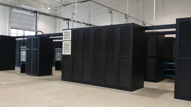 Rows of black louvred server cabinets on a bare concrete floor in a data centre hall, with a roll-up door and exposed ceiling services behind them